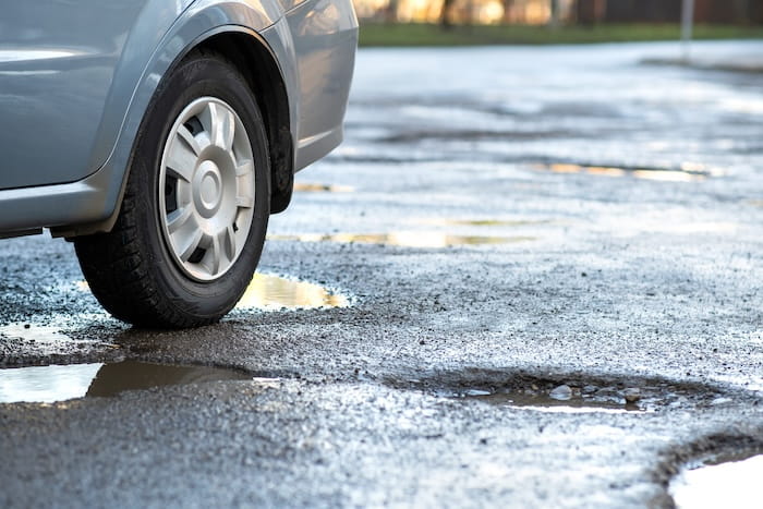 silver car driving through an asphalt driveway that has lots of ruts, puddles, and generally needs maintenance.